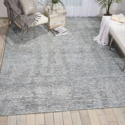 Blue gray woven textured area rug on light wood floor; cozy living room with plants, a metal-framed chair, and a white sofa.