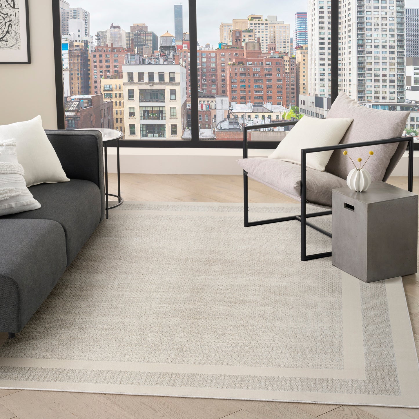 Modern living room with a city-view window, gray sofa & metal-framed chairs, a concrete side table, & beige bordered rug.