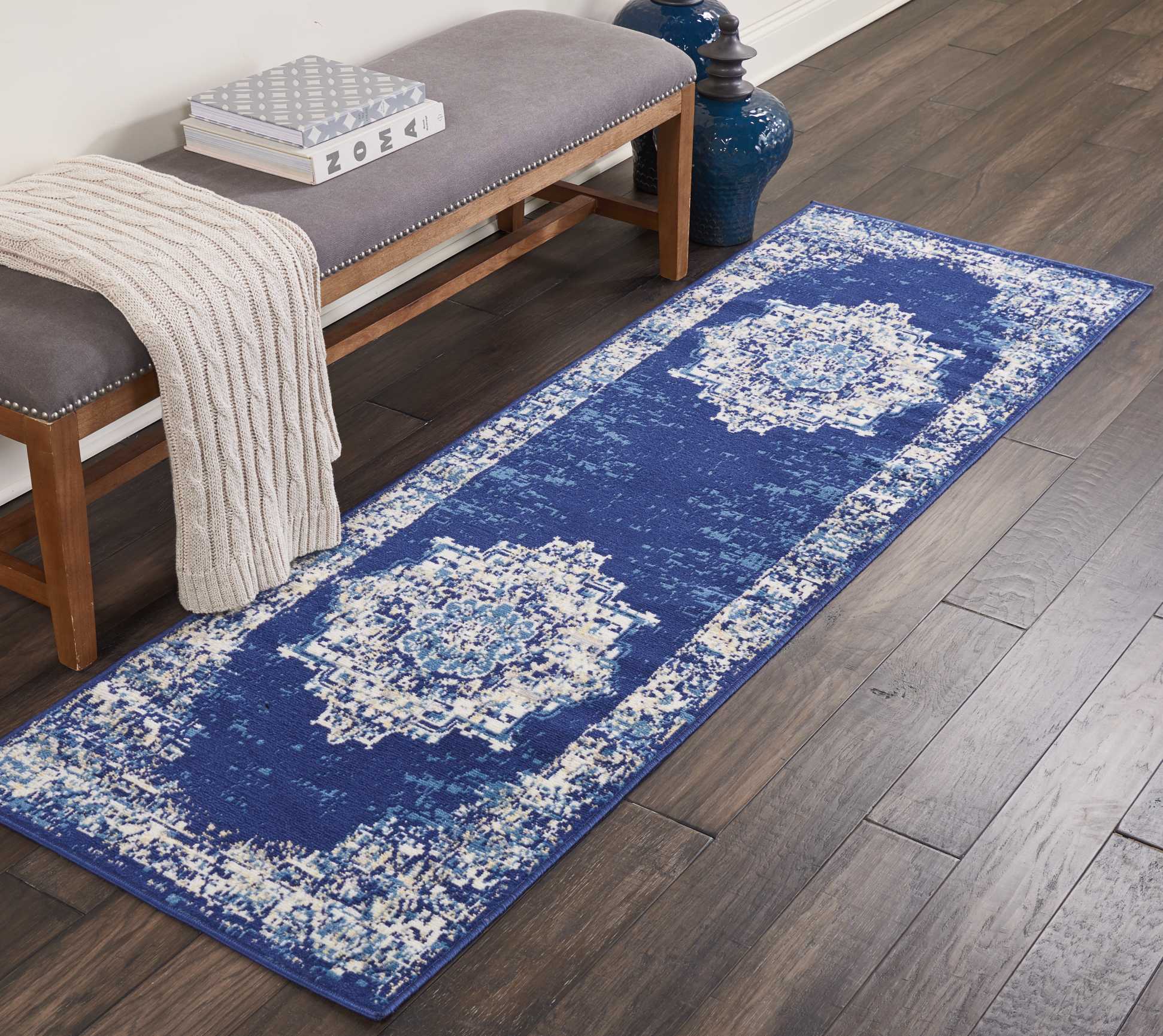 Blue distressed geometric runner rug on dark wood floor beside a wooden bench with gray cushion and beige knit throw.