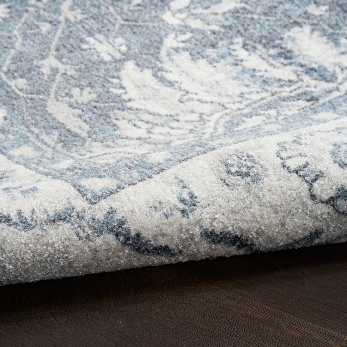 Light blue and ivory patterned area rug with floral motif, folded corner on dark wood floor.