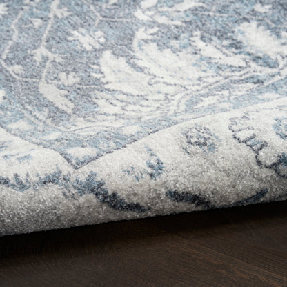 Light blue and ivory patterned area rug with floral motif, folded corner on dark wood floor.