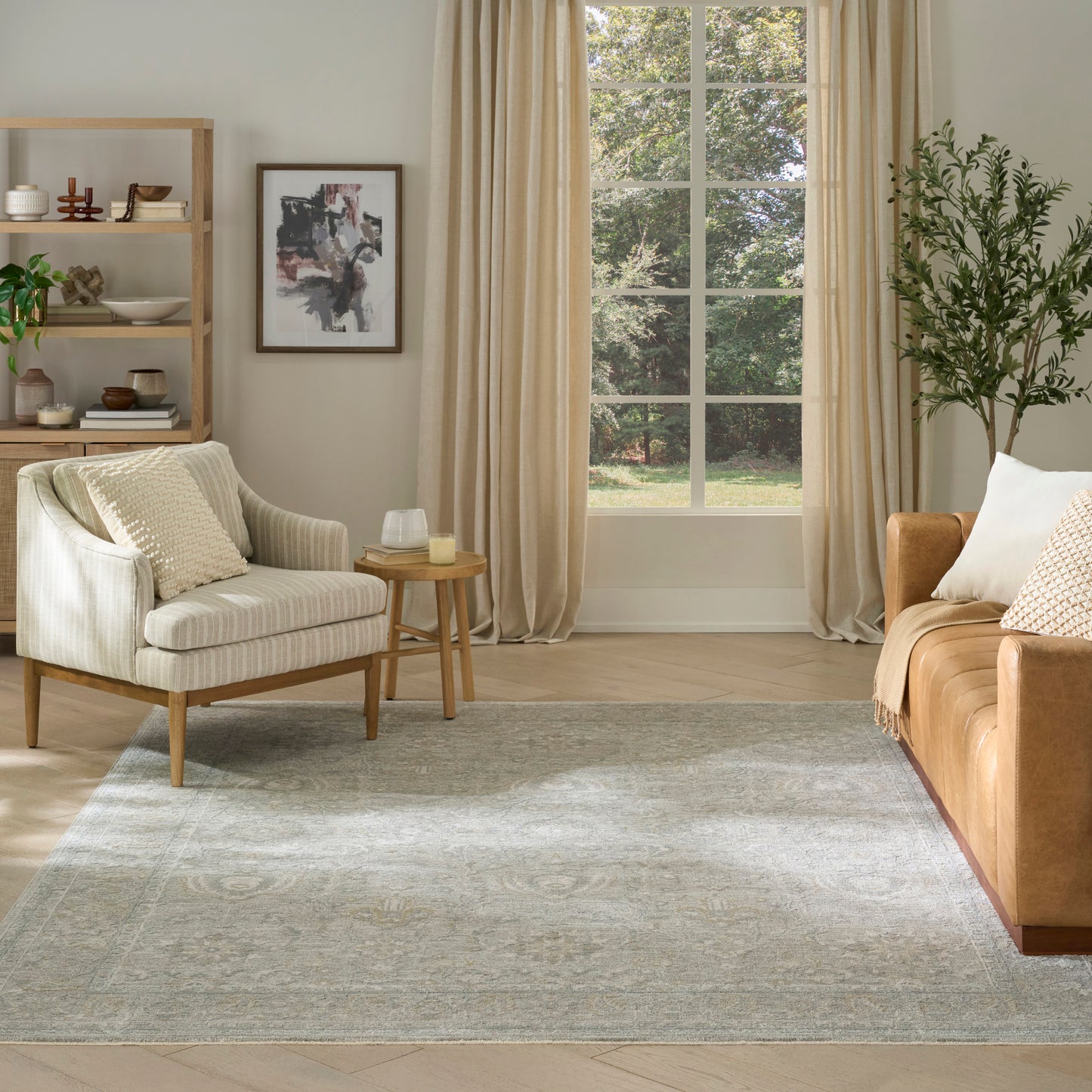 Bright living room with beige armchair, wooden side table, tan sofa, gray rug, bookshelf, tall plant, and cream curtains.