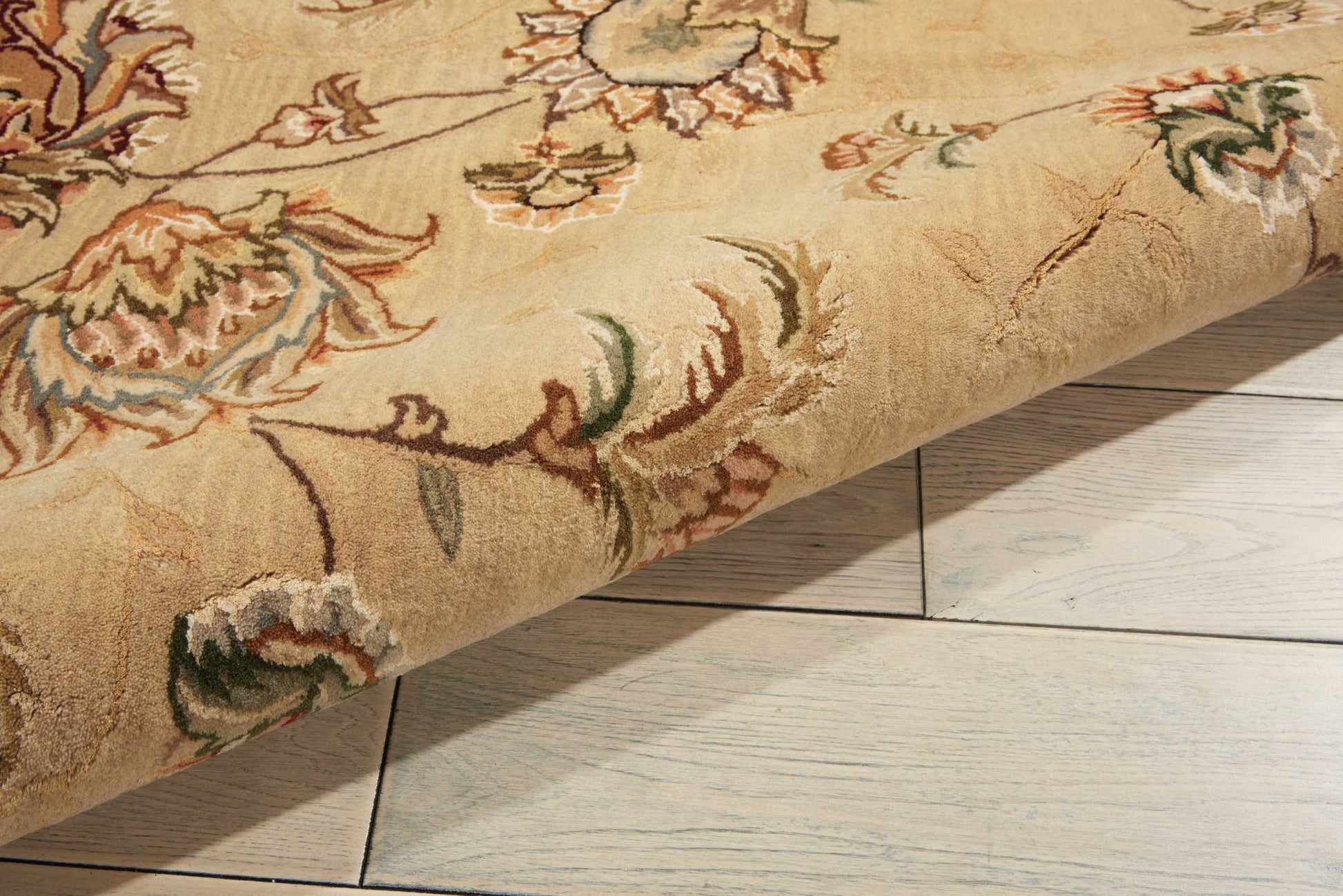 Cream traditional floral rug with tan, brown, green and pink motifs, partially rolled on light wood floor.