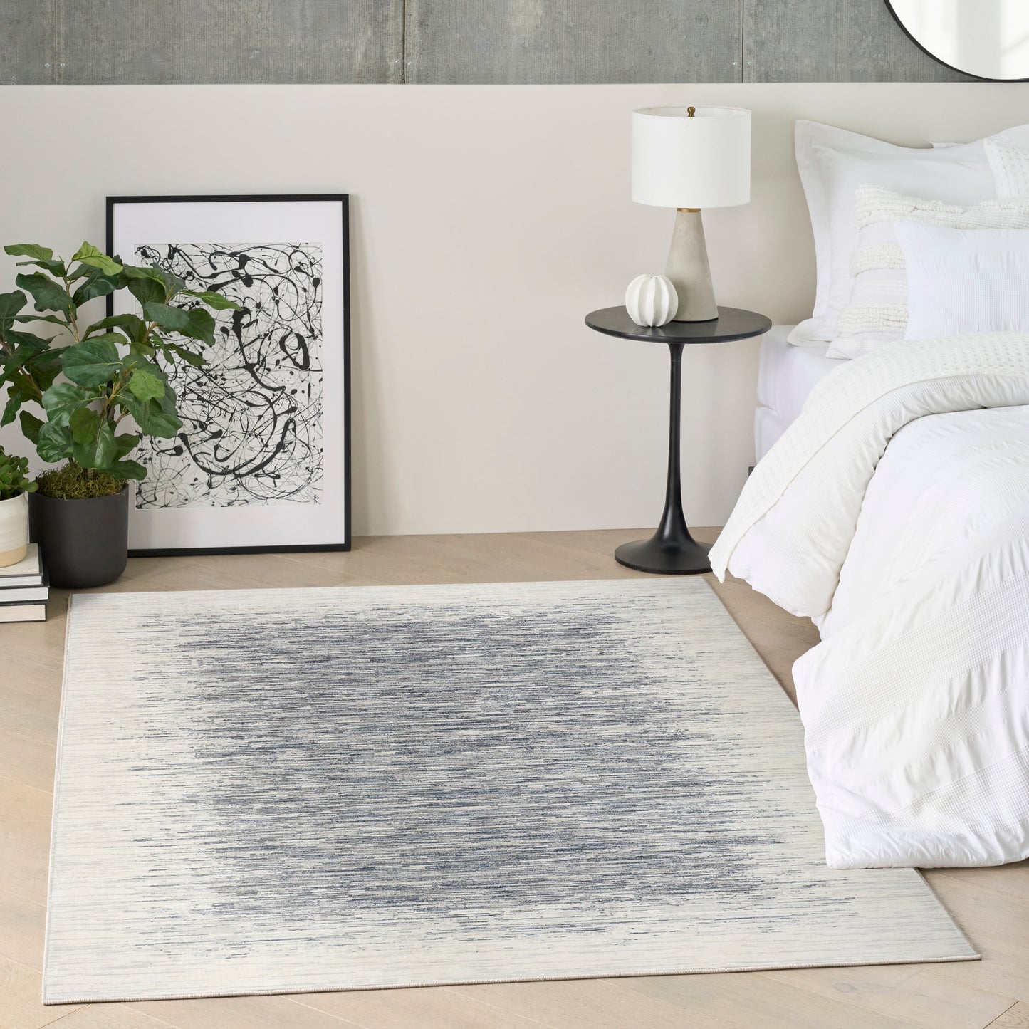 Rectangular flat-weave rug with blue-gray gradient in a modern bedroom with white bedding, black side table, plants, and art.