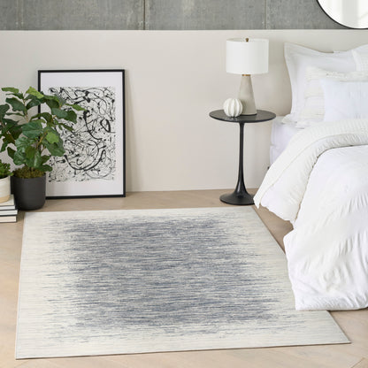 Rectangular flat-weave rug with blue-gray gradient in a modern bedroom with white bedding, black side table, plants, and art.