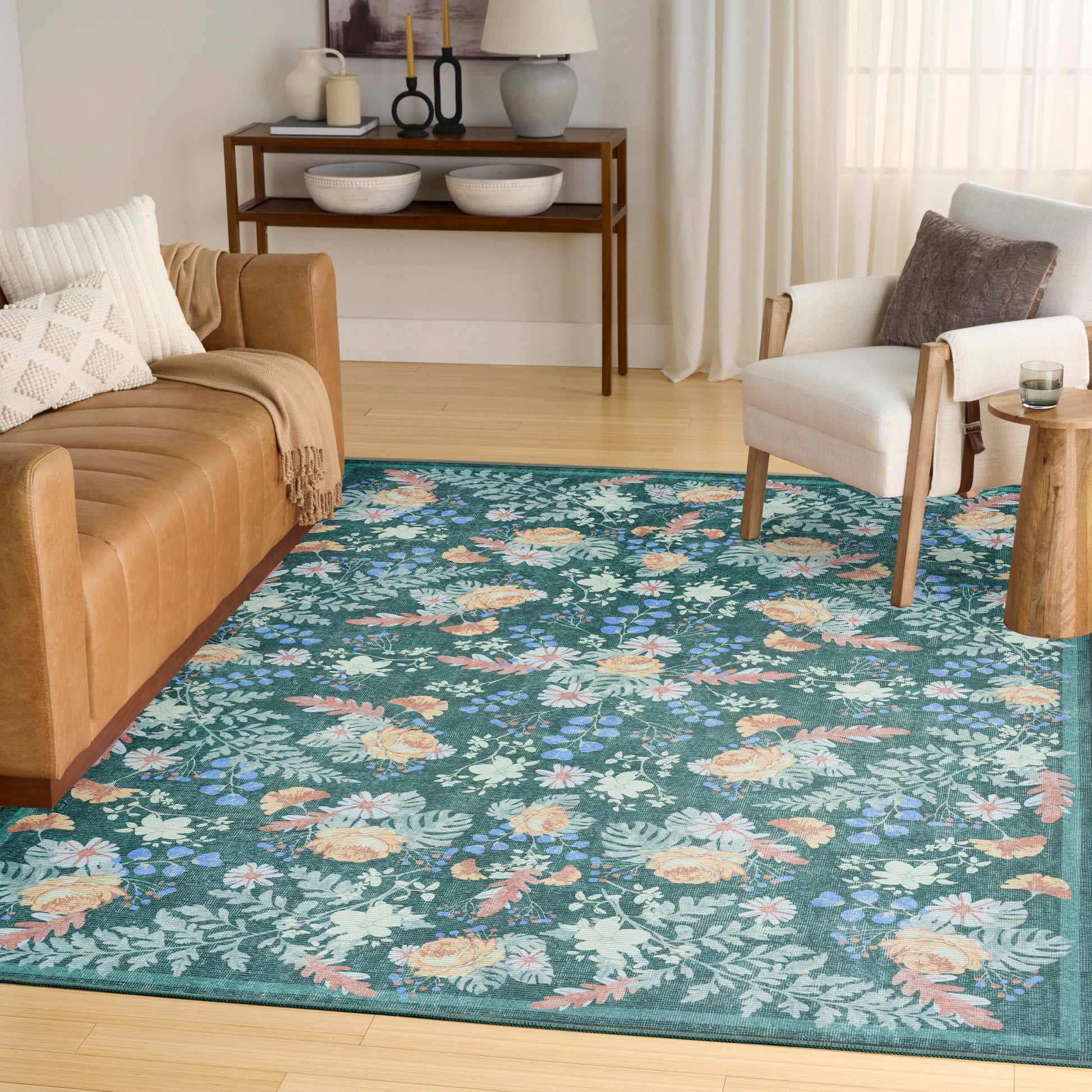 Teal floral area rug on light wood floor with tan leather sofa, white armchair, and wooden console in a bright living room.